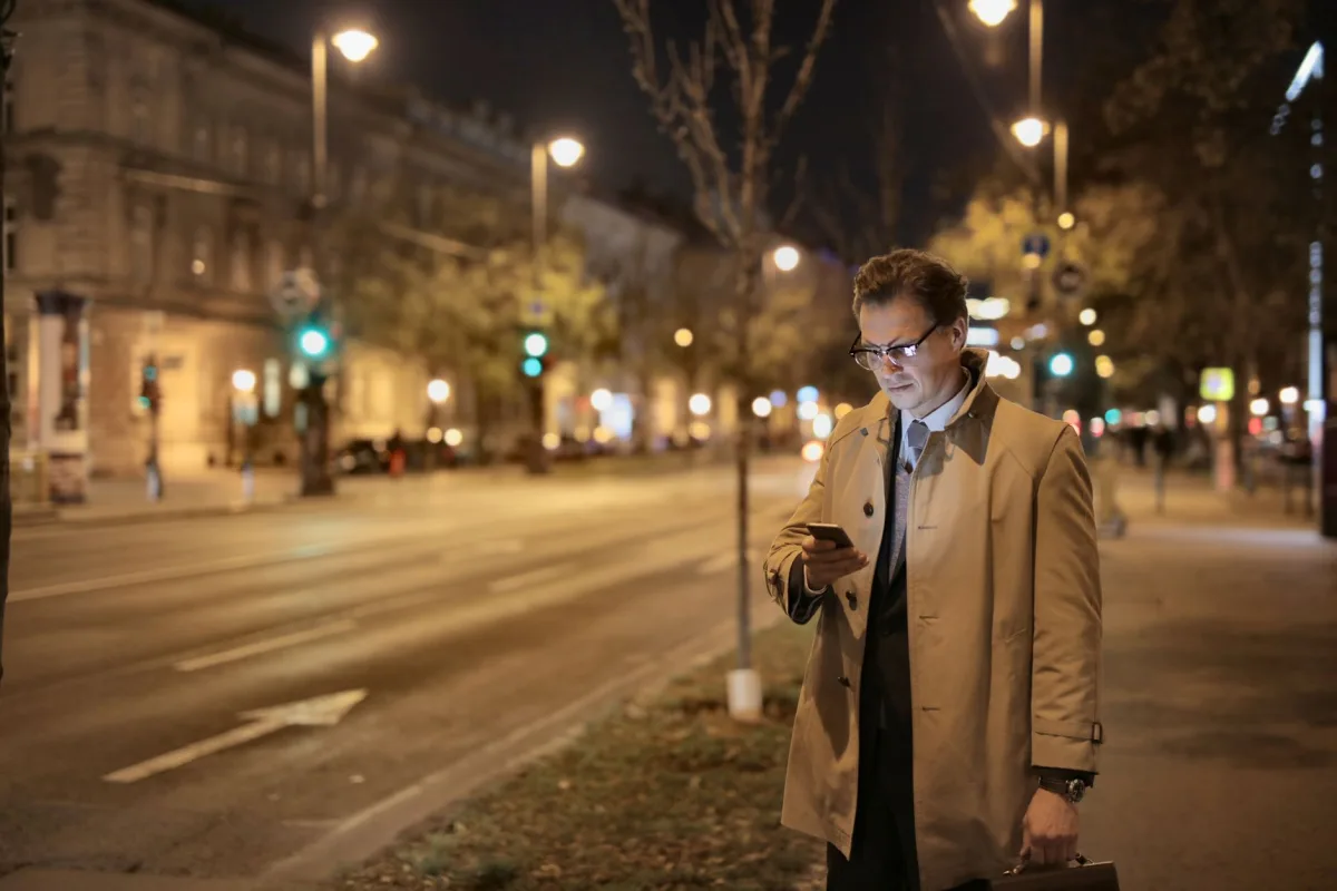 Focused brown haired man browsing mobile phone while standing on empty city street in soft light of streetlamp Focused brown haired man browsing mobile phone while standing on empty city street in soft light of streetlamp