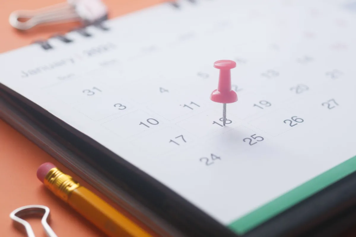 A close-up of a calendar page with a pink push pin, pencil, and binder clip on an orange background.