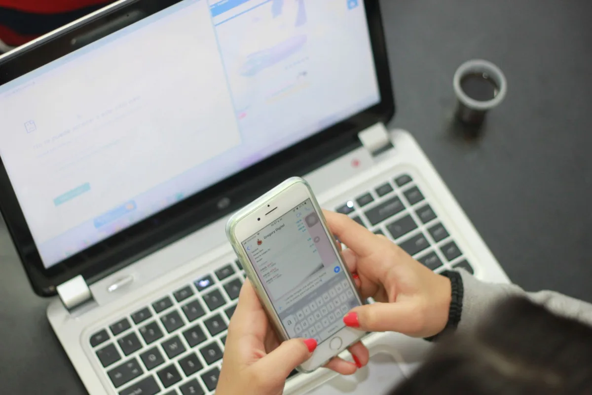 Close-up of woman texting on a smartphone while a laptop is open, indicating multitasking.