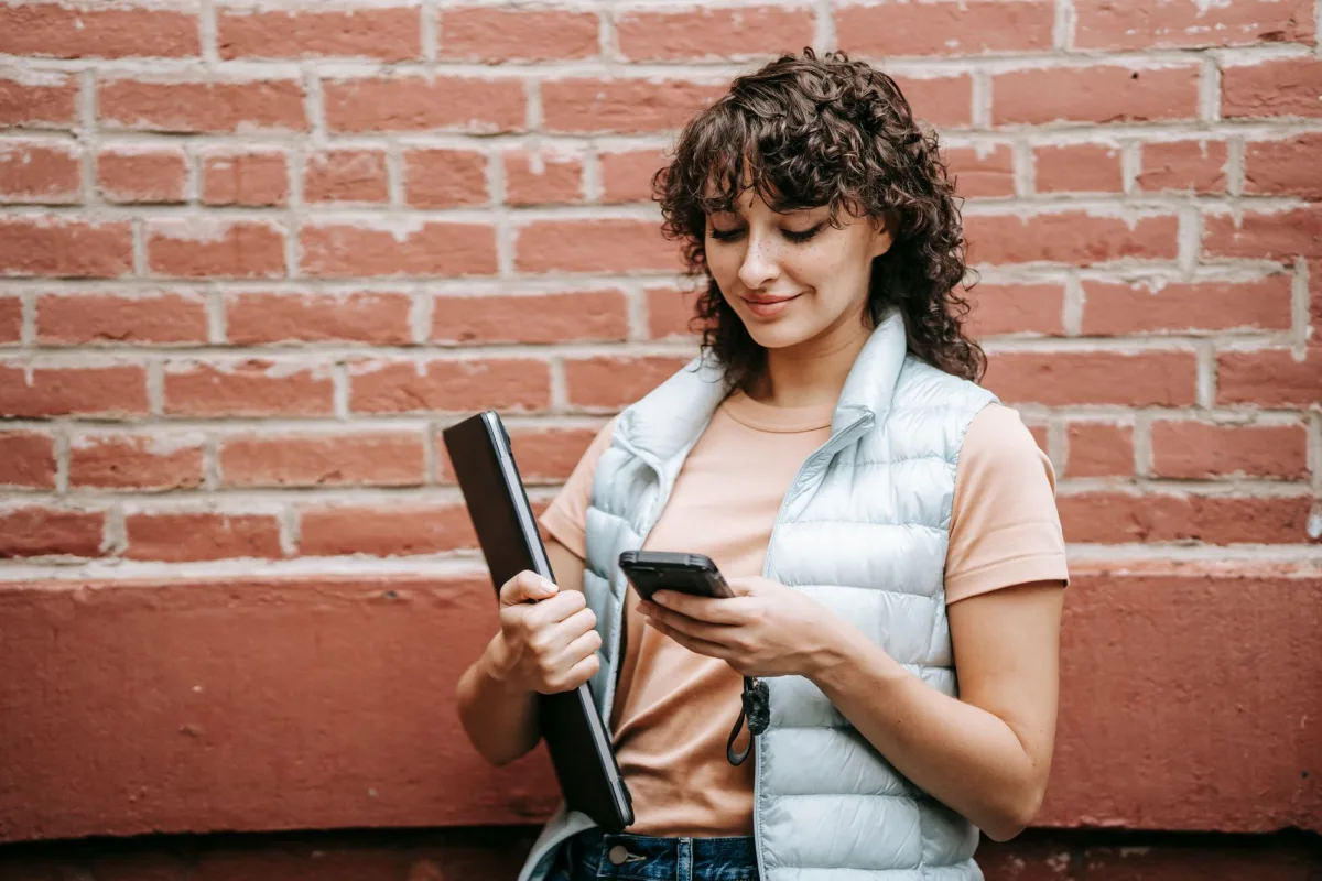 Content adult female remote employee with netbook text messaging on cellphone against brick wall