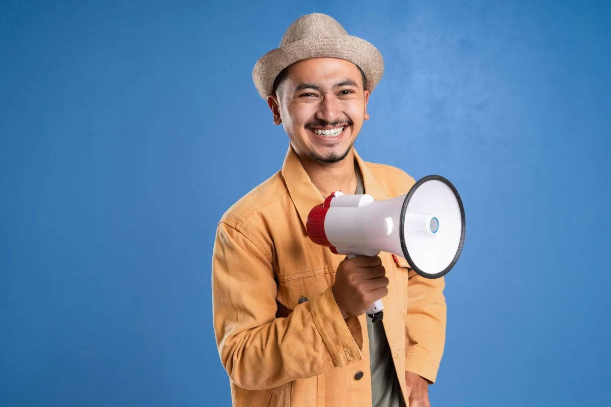 Cheerful man holding a megaphone and wearing a hat, set against a blue background. Cheerful man holding a megaphone and wearing a hat, set against a blue background.