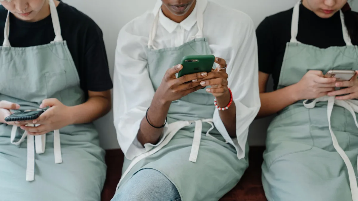 Crop anonymous young diverse waitresses in aprons using smartphones while resting during break at workplace Crop anonymous young diverse waitresses in aprons using smartphones while resting during break at workplace