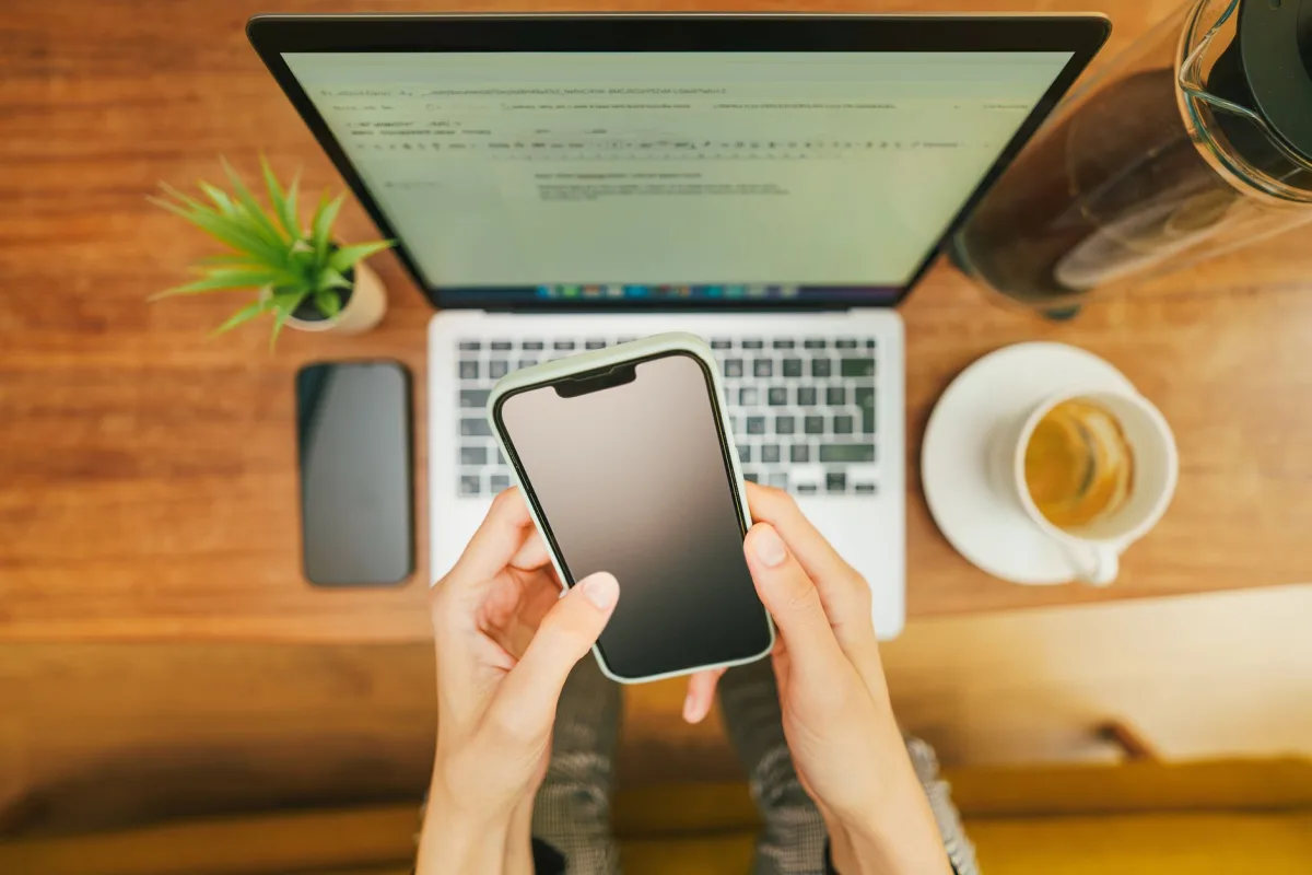Hands holding a smartphone over a laptop with coffee in a cozy home office setup. Hands holding a smartphone over a laptop with coffee in a cozy home office setup.