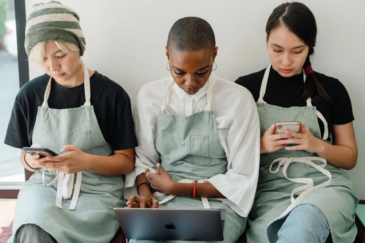 Focused young multiethnic female colleagues in casual clothes and aprons sitting and leaning on white wall while using gadgets