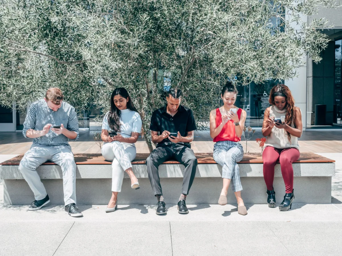 Group of diverse adults sitting outdoors using smartphones under a tree. Group of diverse adults sitting outdoors using smartphones under a tree.