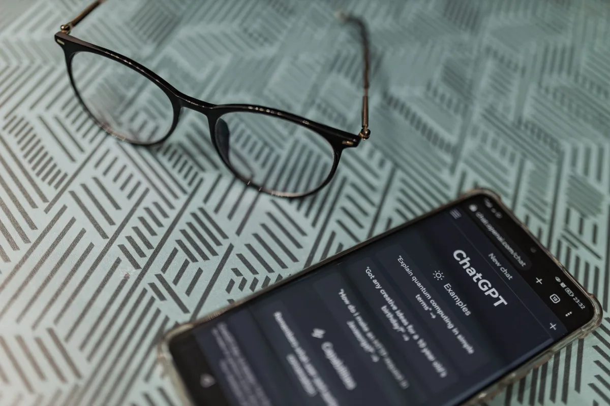 Eyeglasses next to a smartphone displaying the ChatGPT app on a patterned surface. Eyeglasses next to a smartphone displaying the ChatGPT app on a patterned surface.
