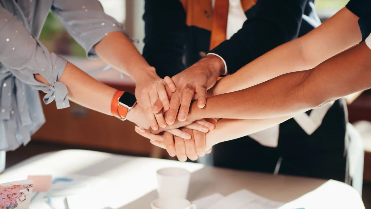 A close-up shot of diverse team members stacking hands over a white table, symbolizing unity and collaboration. A close-up shot of diverse team members stacking hands over a white table, symbolizing unity and collaboration.