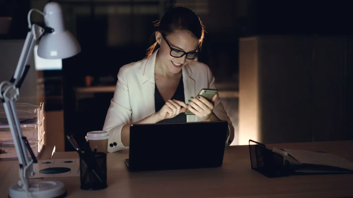 Smiling businesswoman working late, checking smartphone in office.
