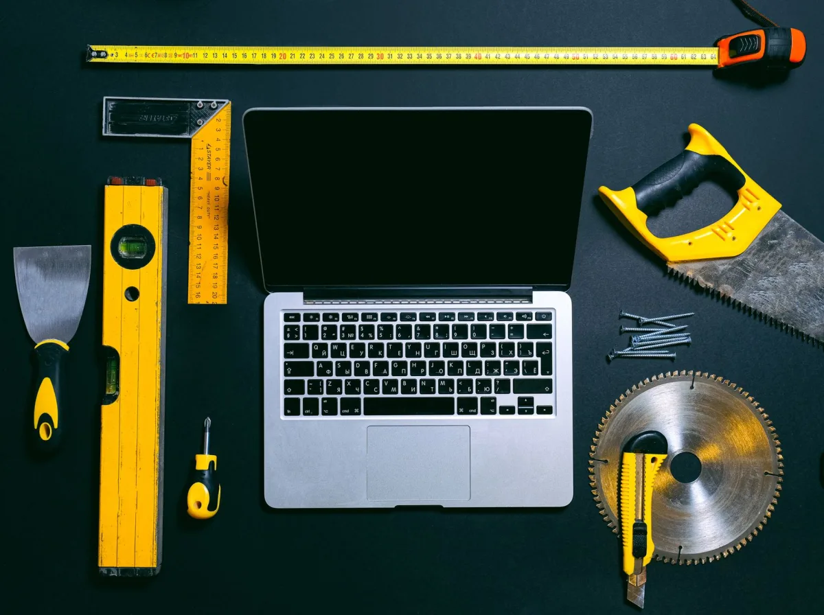 Overhead view of a laptop surrounded by various hand tools on a dark background, representing technology and construction. Overhead view of a laptop surrounded by various hand tools on a dark background, representing technology and construction.
