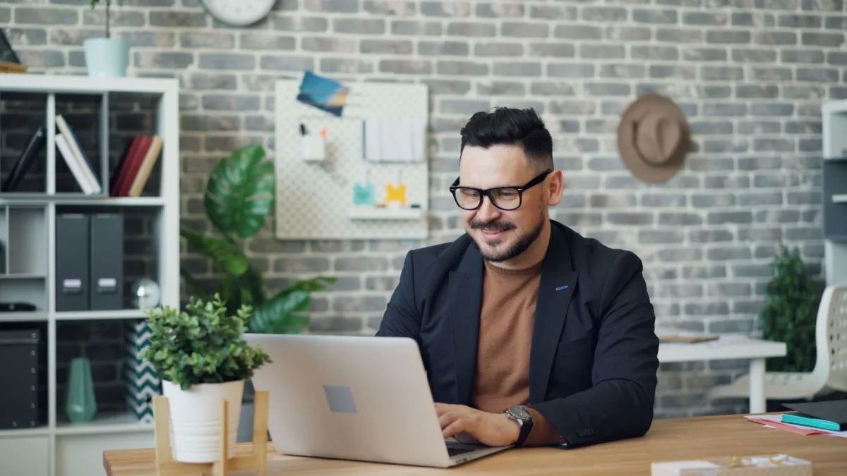A man smiles while working on a laptop in a stylish, modern office setting. A man smiles while working on a laptop in a stylish, modern office setting.