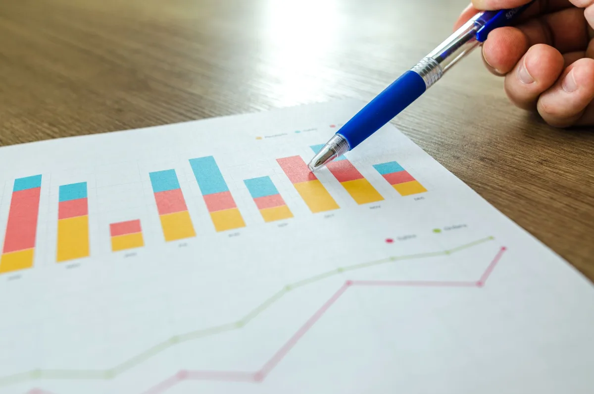 Detailed close-up of a hand pointing at colorful charts with a blue pen on wooden surface. Detailed close-up of a hand pointing at colorful charts with a blue pen on wooden surface.