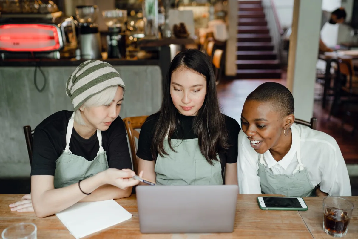 Optimistic smiling diverse female colleagues in aprons browsing netbook and preparing for exam while sitting at table in cozy cafeteria Optimistic smiling diverse female colleagues in aprons browsing netbook and preparing for exam while sitting at table in cozy cafeteria