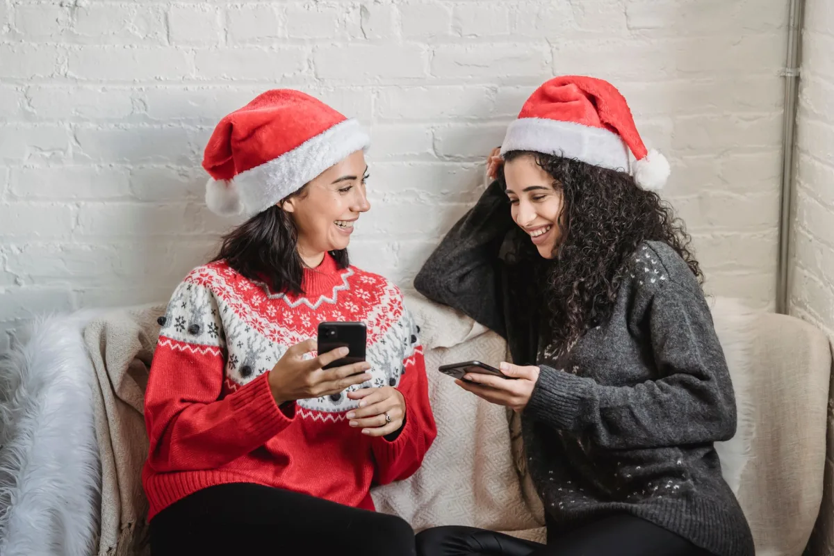 Cheerful young Hispanic female friends in warm jumpers and Santa hats sitting on small sofa and browsing smartphones while spending Christmas holiday together Cheerful young Hispanic female friends in warm jumpers and Santa hats sitting on small sofa and browsing smartphones while spending Christmas holiday together