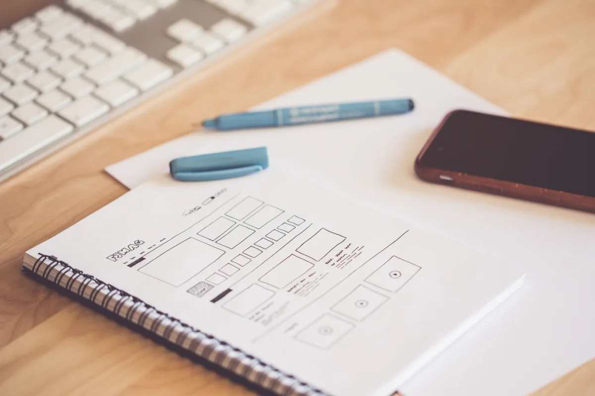 A neat workspace featuring a sketchpad with wireframes, a smartphone, and a keyboard on a wooden desk. A neat workspace featuring a sketchpad with wireframes, a smartphone, and a keyboard on a wooden desk.