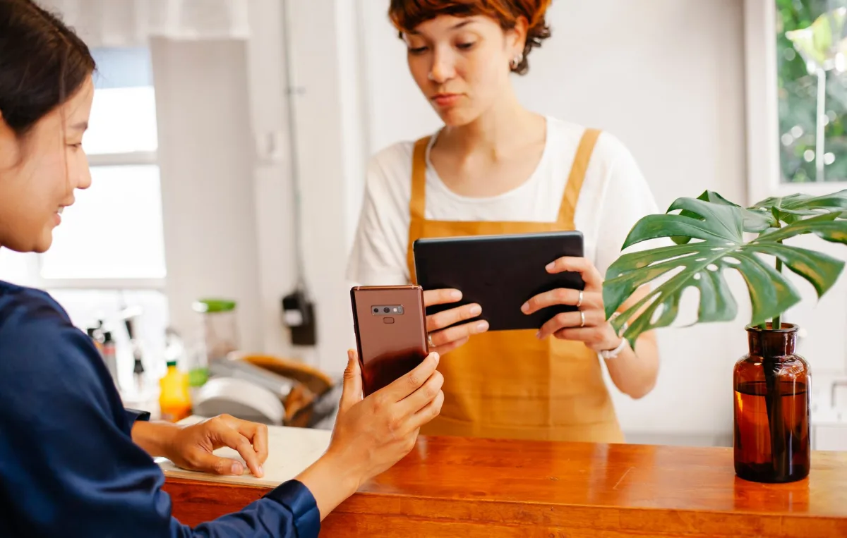Crop multiethnic female partners with cellphone and tablet interacting at cafeteria counter with Monstera sprig in vase Crop multiethnic female partners with cellphone and tablet interacting at cafeteria counter with Monstera sprig in vase