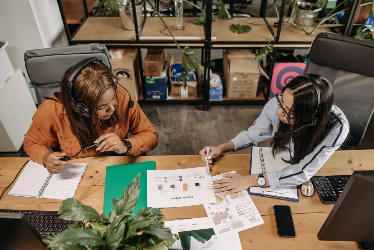 Two women wearing headsets discuss work at a shared desk in an office. Two women wearing headsets discuss work at a shared desk in an office.