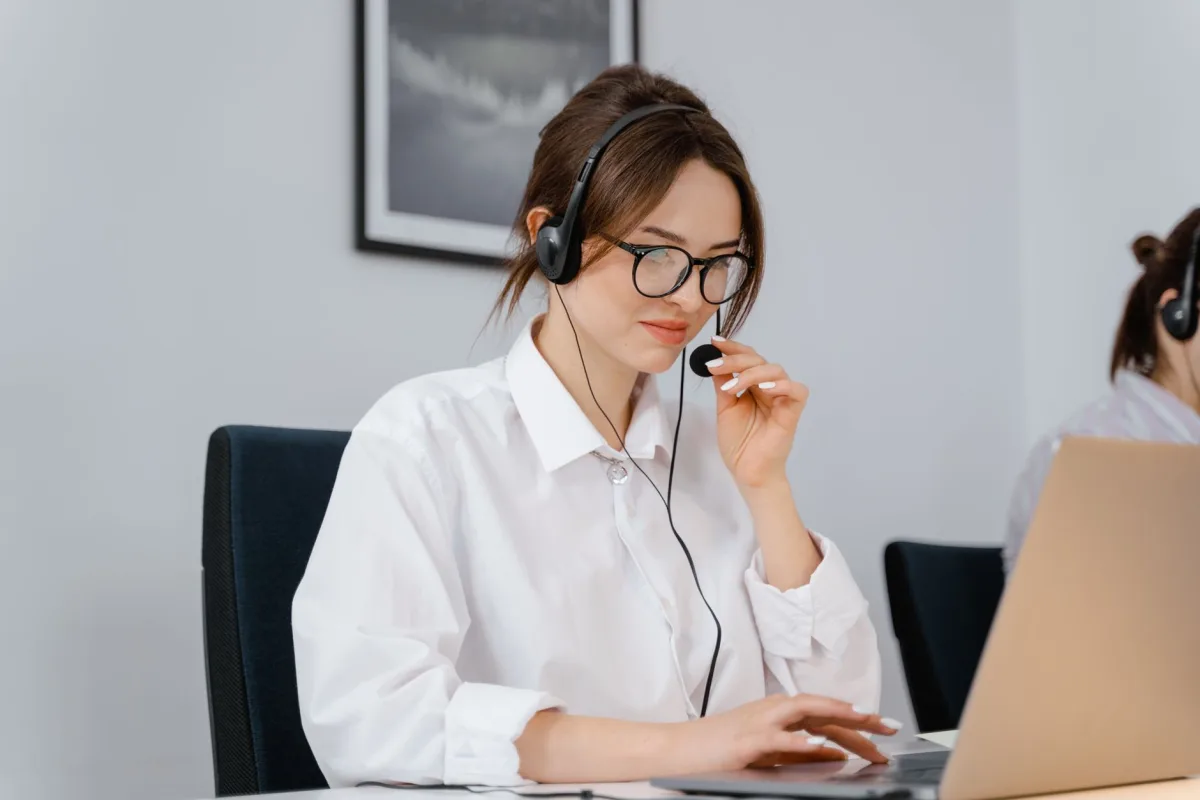 Young woman in glasses and headset providing support at a laptop in an office setting.