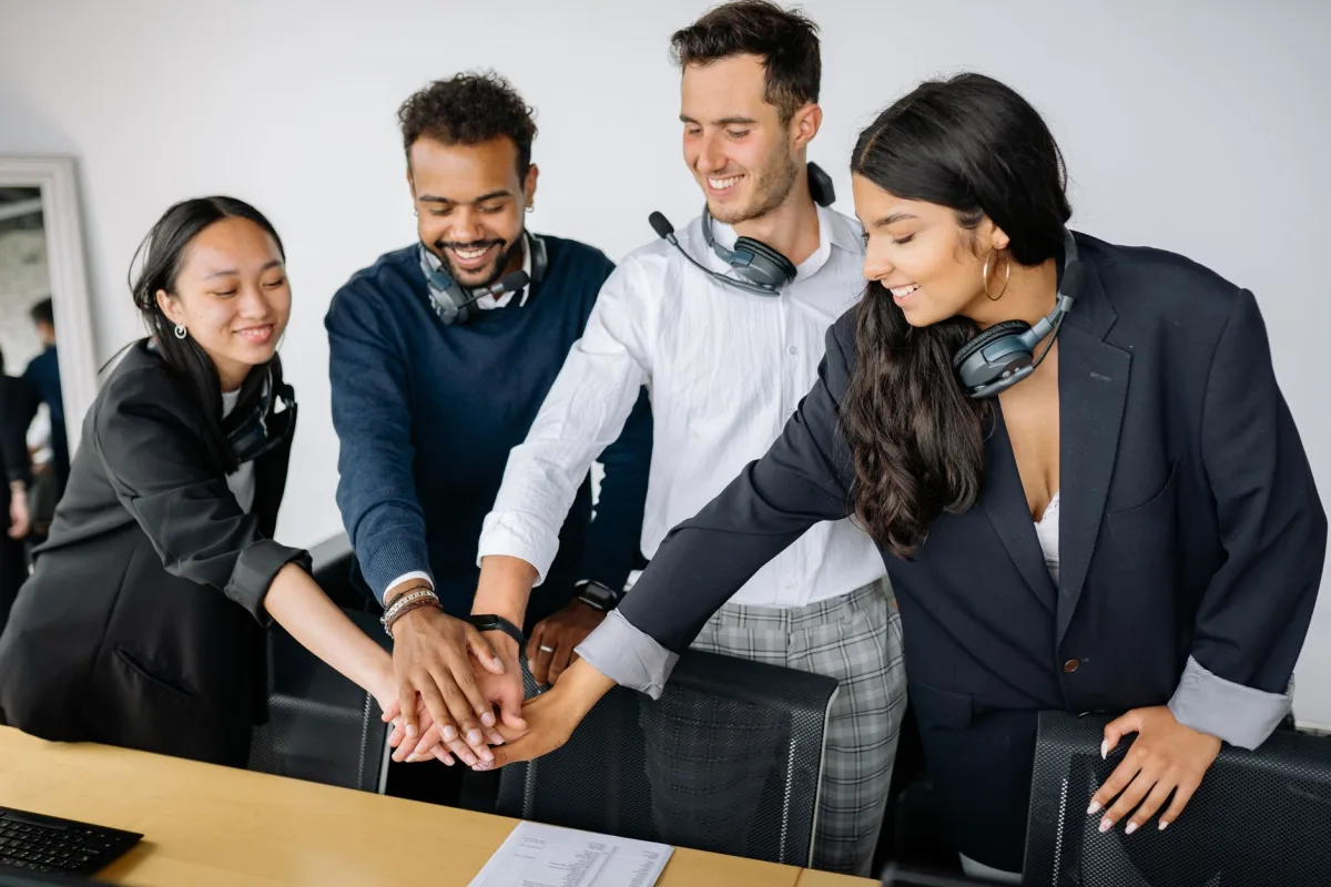 A diverse group of call center agents in an office joining hands in a teamwork gesture, smiling and wearing headsets. A diverse group of call center agents in an office joining hands in a teamwork gesture, smiling and wearing headsets.