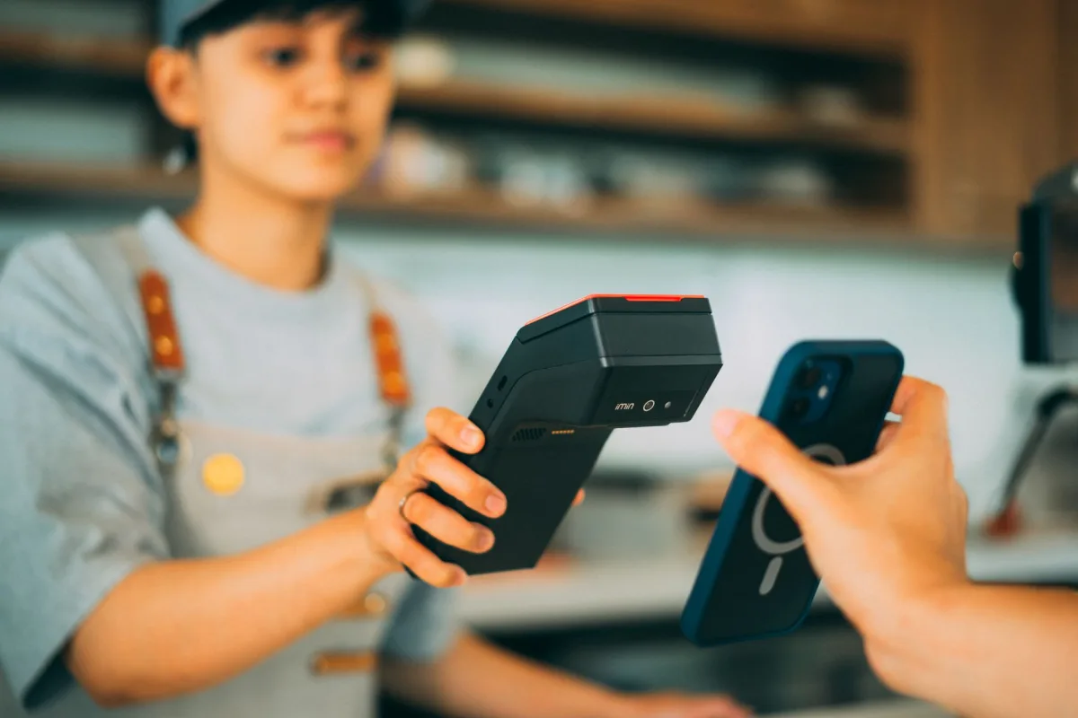 A cashier processes a contactless payment using a smartphone and card reader. A cashier processes a contactless payment using a smartphone and card reader.