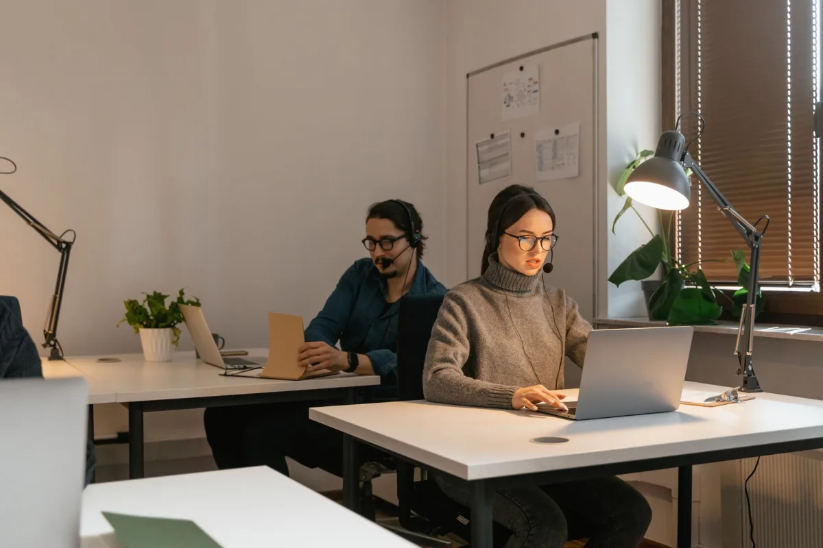 Two colleagues working in an office setting, focusing on laptops and customer
