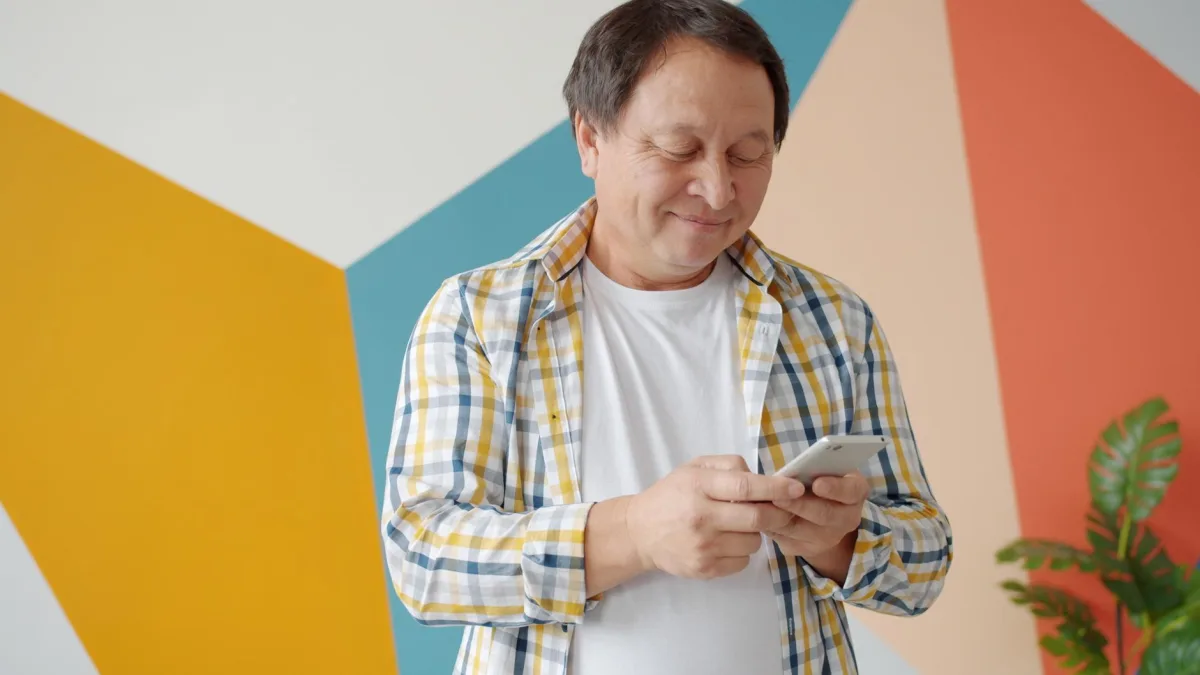 Happy senior man texting on smartphone against colorful wall indoors.