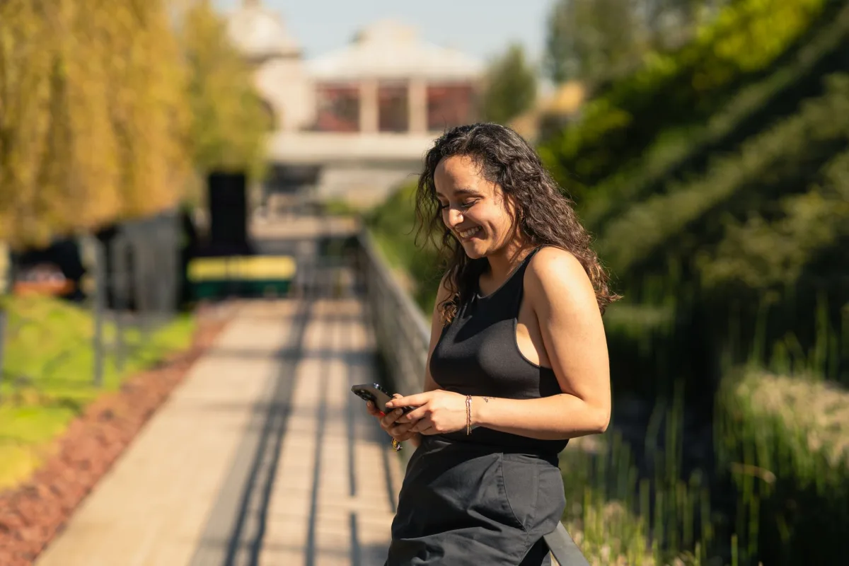 A young woman smiles while texting on her phone in a sunny park setting. A young woman smiles while texting on her phone in a sunny park setting.