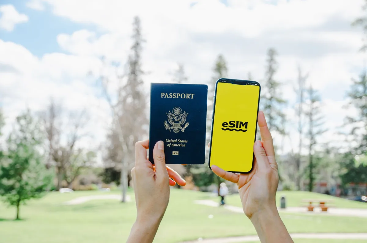 Hands holding a USA passport and smartphone with eSIM in a sunny park.