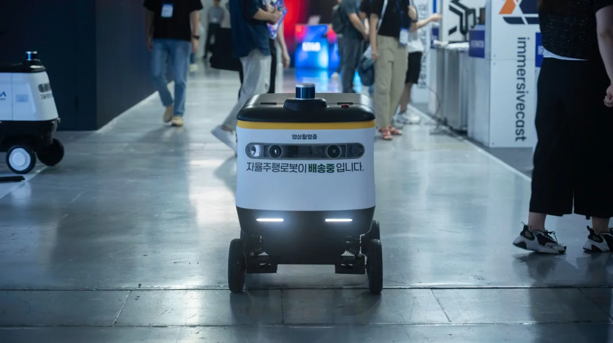 Autonomous delivery robot navigating indoors during a technology event. Autonomous delivery robot navigating indoors during a technology event.