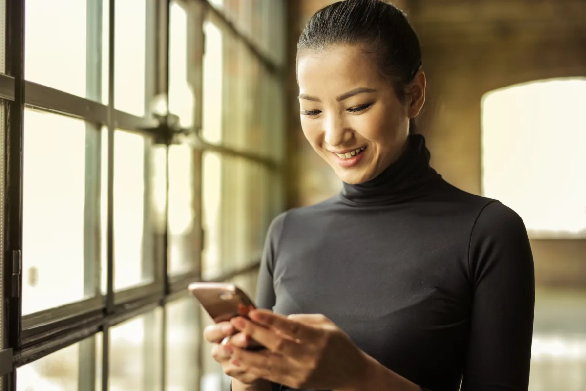 Happy woman in black turtleneck uses smartphone by a sunlit window indoors. Happy woman in black turtleneck uses smartphone by a sunlit window indoors.