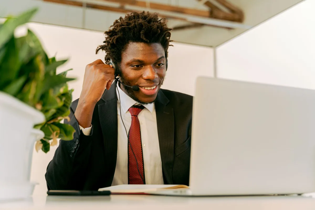 Happy businessman with afro hair wearing a headset and suit, working on a laptop in an office.