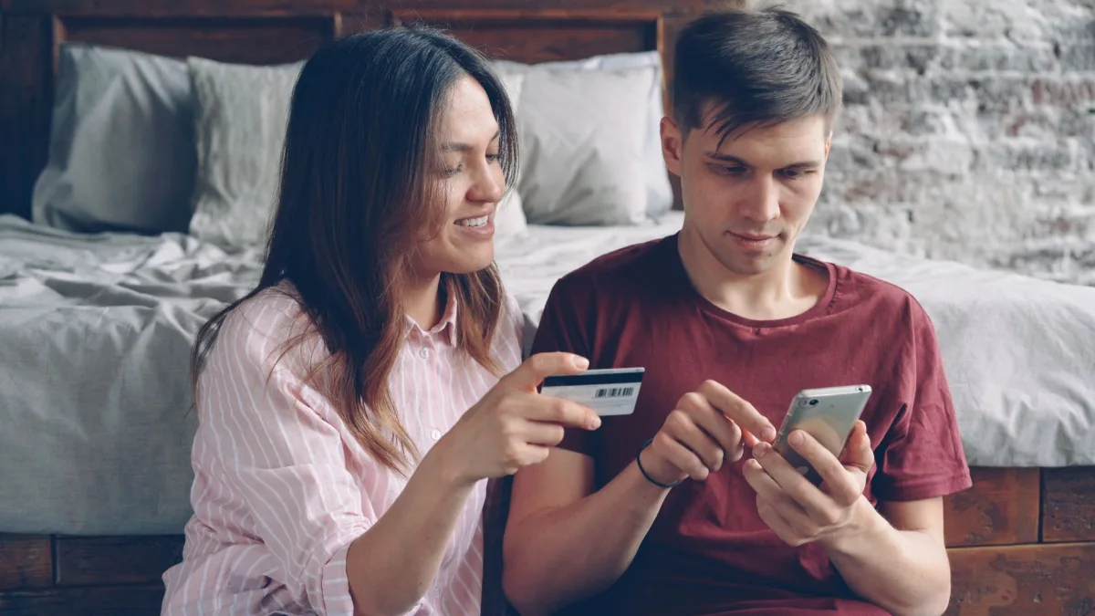 A couple sits on a bed shopping online using a smartphone and credit card in a cozy indoor setting.
