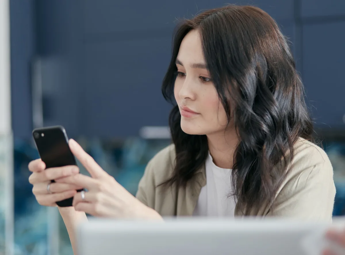 A young woman with black hair using a smartphone indoors, focused on the screen.