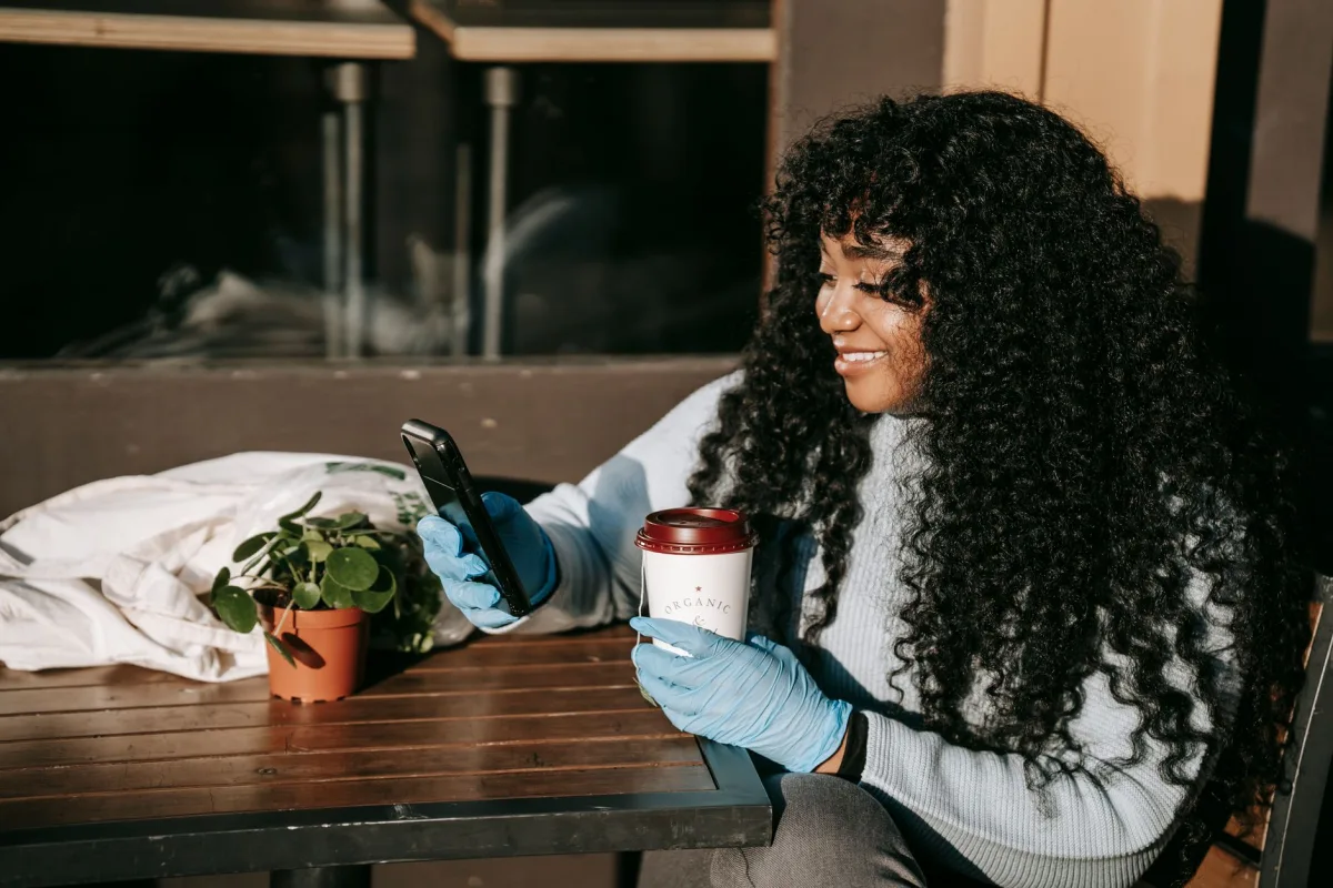 Positive black lady in protective gloves sitting at wooden table with paper cup of coffee near potted green plant while surfing on phone in sunny day in street near building Positive black lady in protective gloves sitting at wooden table with paper cup of coffee near potted green plant while surfing on phone in sunny day in street near building