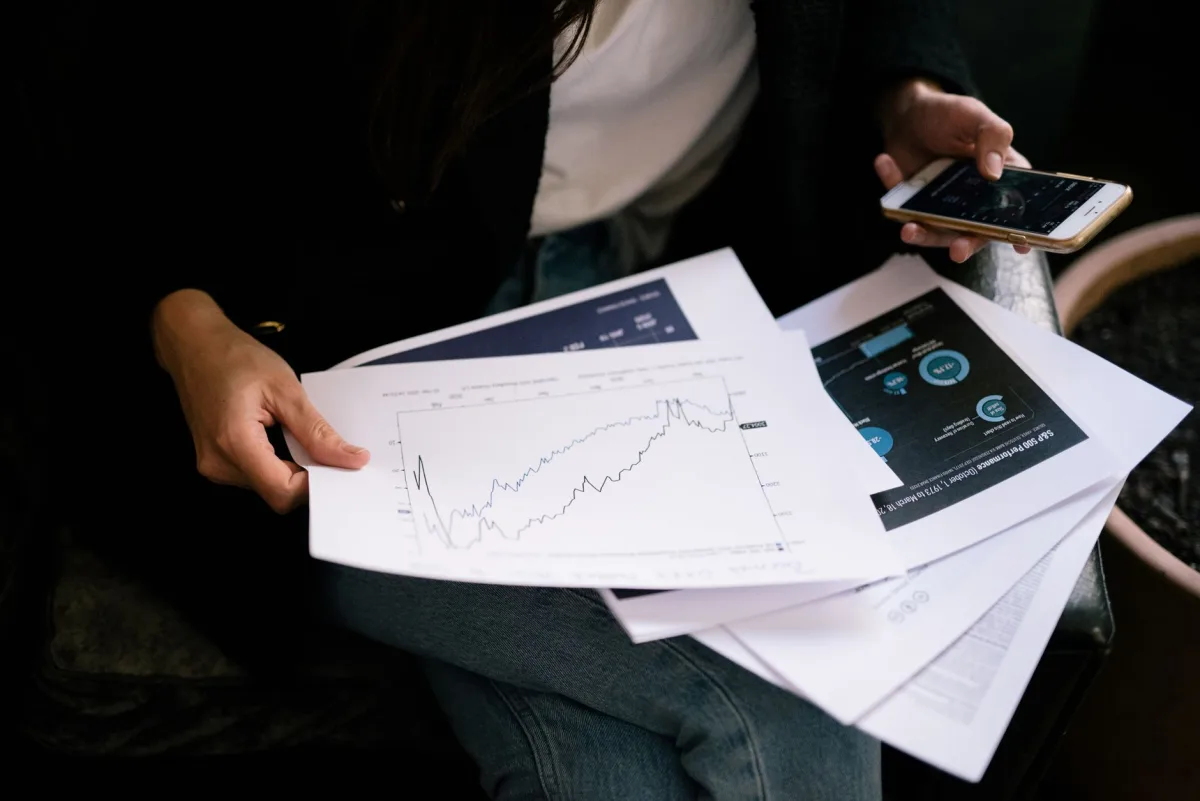 A person reviews stock market graphs on paper and smartphone for business insights. A person reviews stock market graphs on paper and smartphone for business insights.