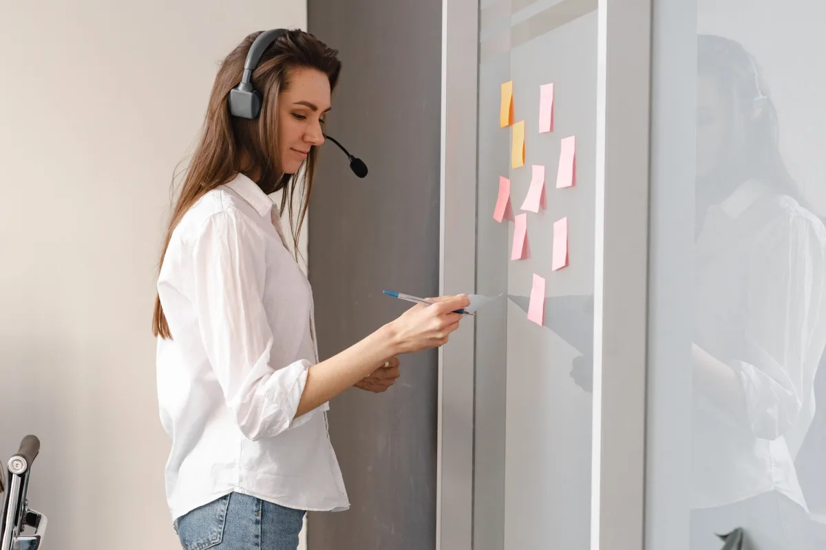 Woman wearing headset organizing tasks on a glass board with sticky notes in an office setting. Woman wearing headset organizing tasks on a glass board with sticky notes in an office setting.