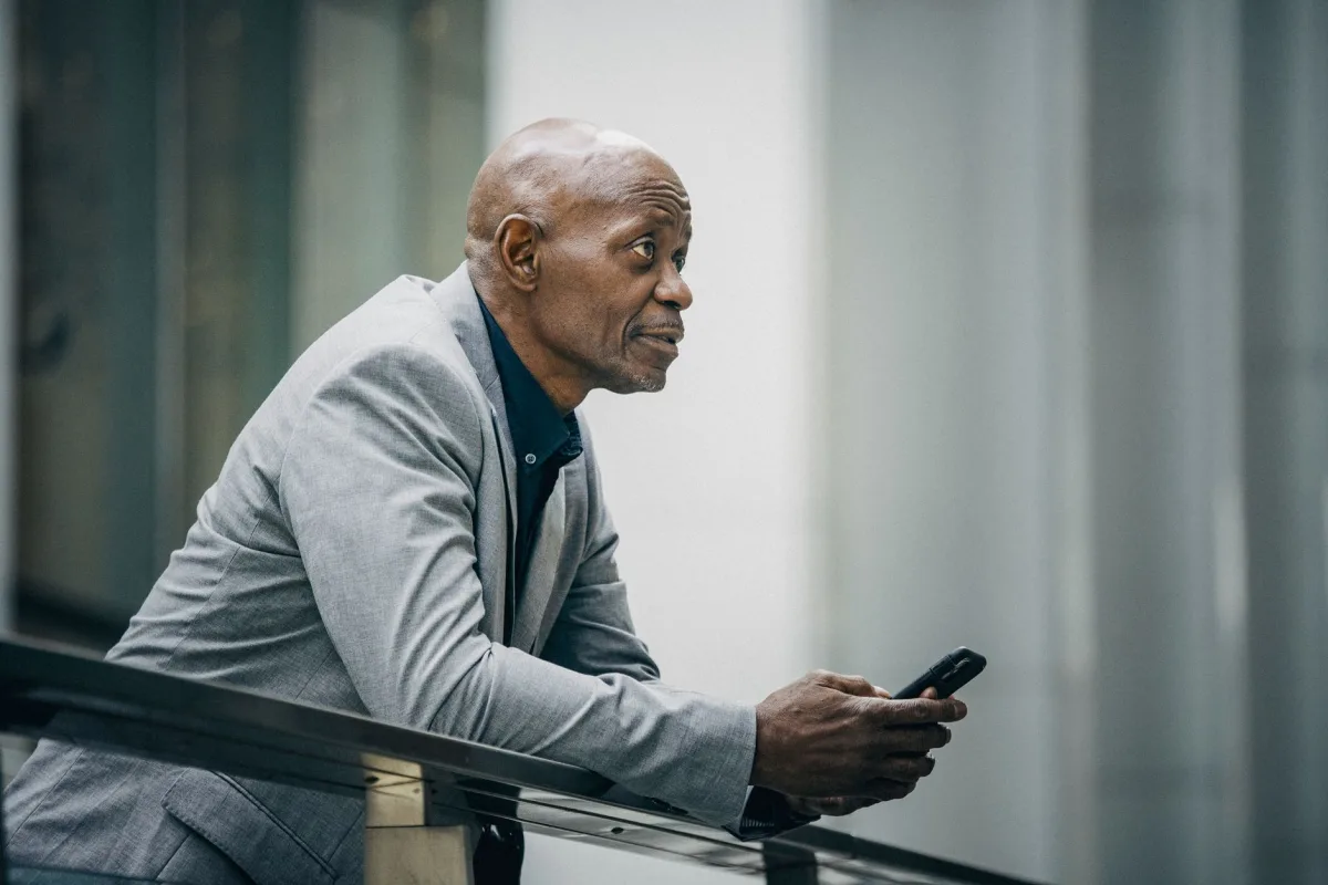 Side view of concentrated bald African American man leaning on fence while using phone and looking away in modern city in soft daylight Side view of concentrated bald African American man leaning on fence while using phone and looking away in modern city in soft daylight