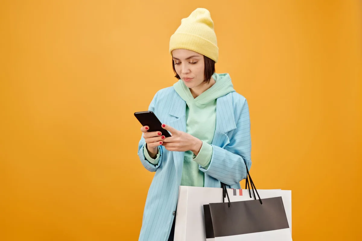 Trendy young woman using smartphone and holding shopping bags against yellow background.