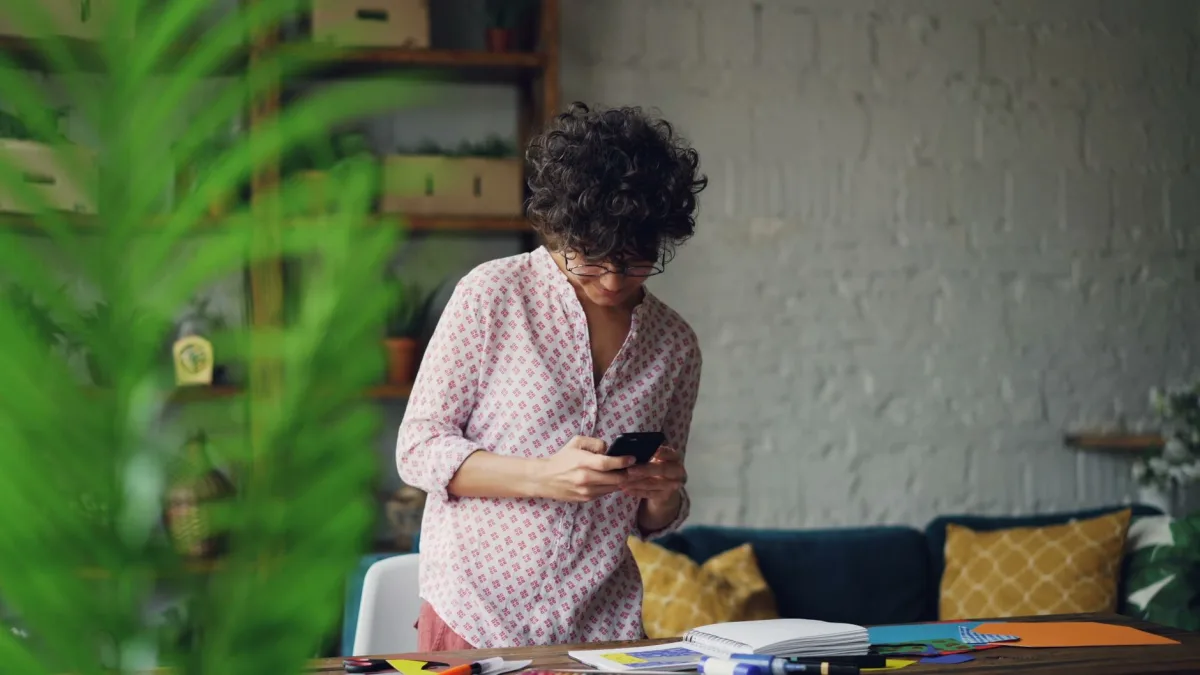 A young woman in a home office browses her smartphone, surrounded by plants and notebooks. A young woman in a home office browses her smartphone, surrounded by plants and notebooks.