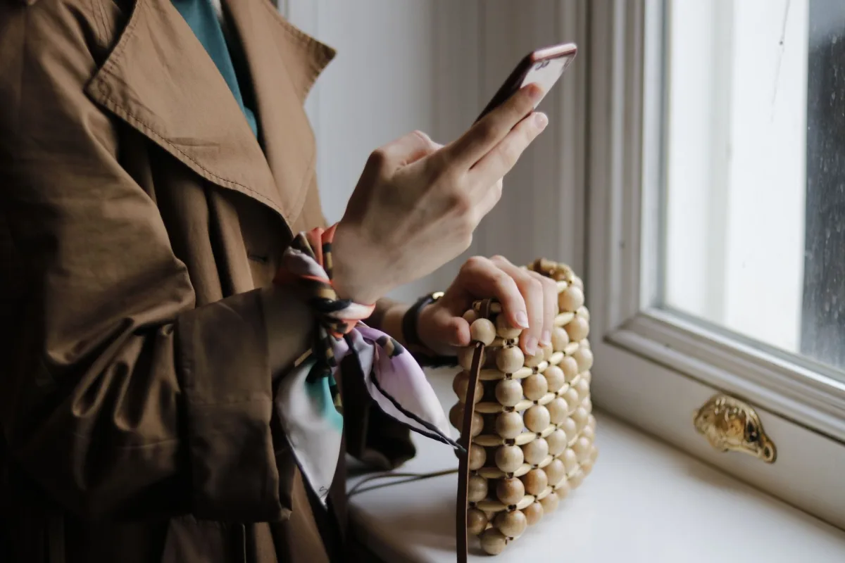 Crop unrecognizable female with trendy handbag text on cellphone above windowsill in building in daytime Crop unrecognizable female with trendy handbag text on cellphone above windowsill in building in daytime