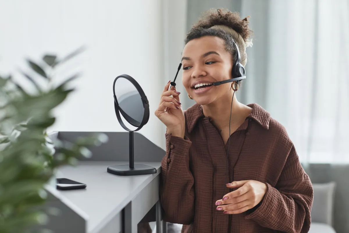 Young woman applying makeup while wearing a headset, smiling in front of a mirror indoors.