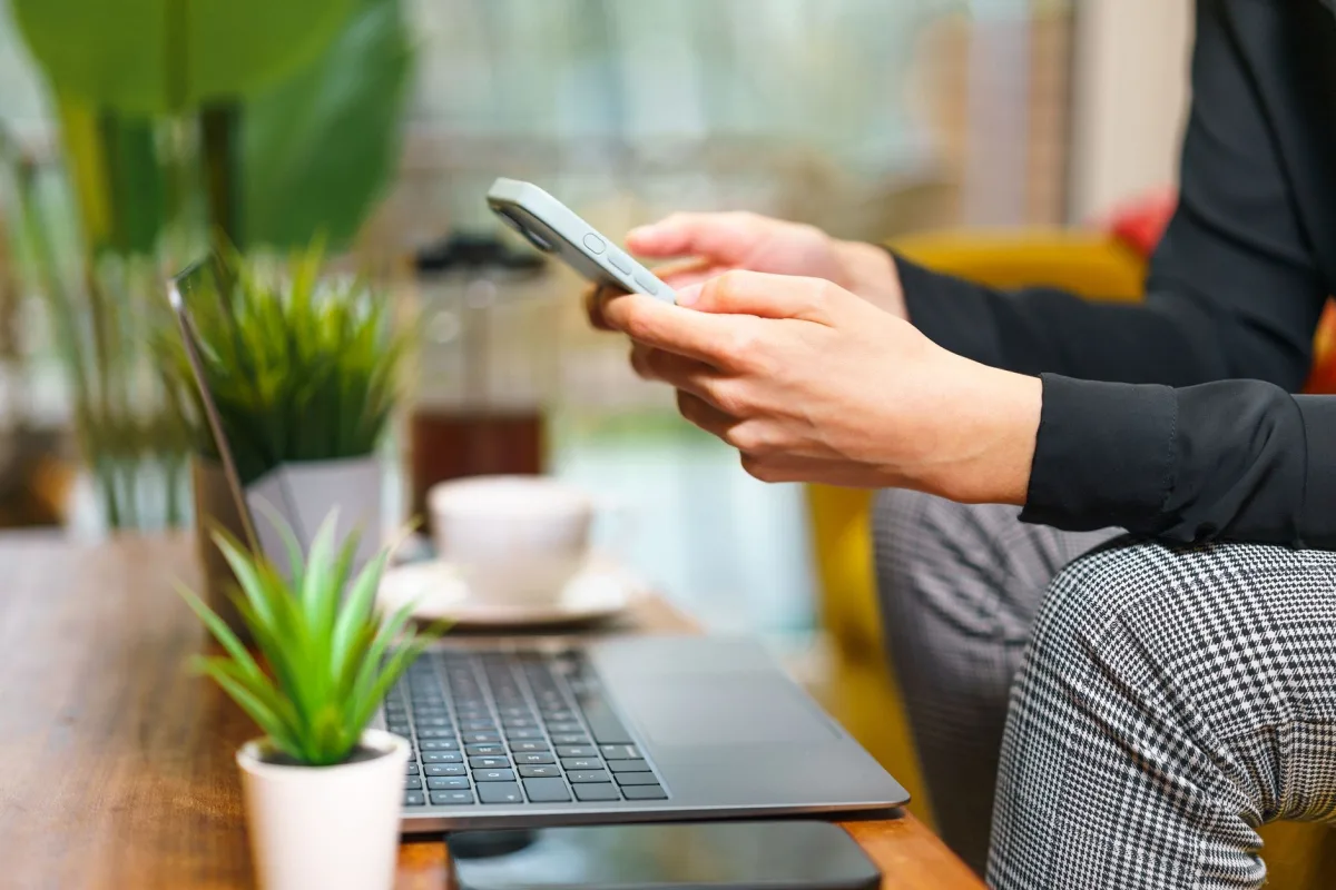 A person using a smartphone in a cozy home office setting with plants, a laptop, and coffee. A person using a smartphone in a cozy home office setting with plants, a laptop, and coffee.