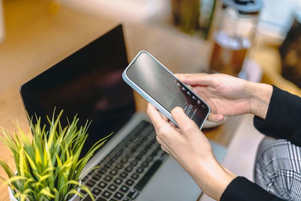 A woman holding a smartphone above a laptop in a cozy home office setting. A woman holding a smartphone above a laptop in a cozy home office setting.