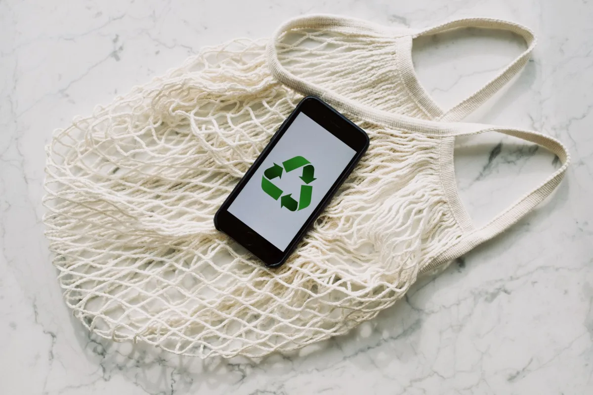 Top view of smartphone with recycling sign on screen placed on white textile mesh bag on white marble table in light room Top view of smartphone with recycling sign on screen placed on white textile mesh bag on white marble table in light room