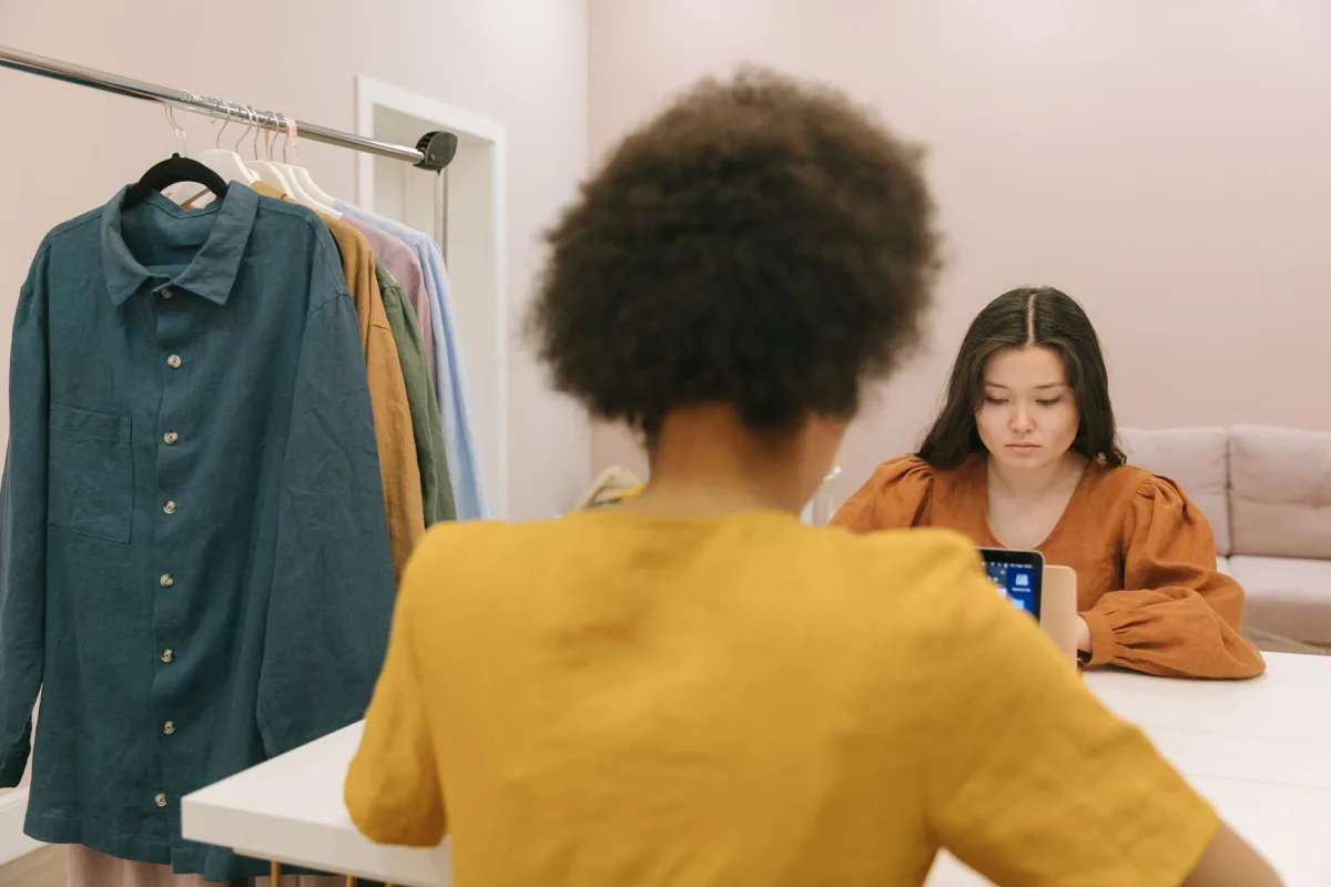 Two women working in a styled workspace with clothes rack and device focus. Two women working in a styled workspace with clothes rack and device focus.