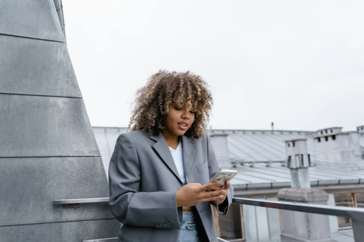 A young woman with curly hair texting on her smartphone while on a rooftop.