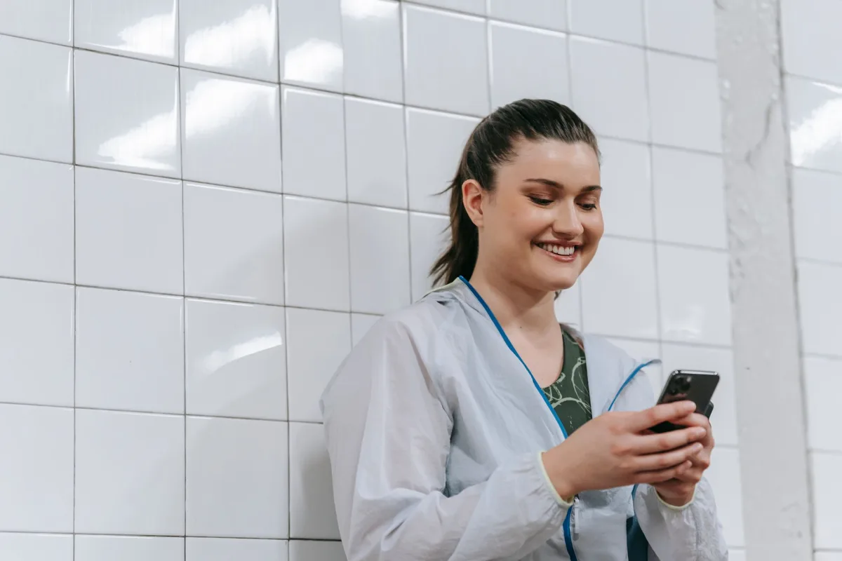 A woman happily texts on her smartphone against a white tiled wall indoors, wearing a casual jacket. A woman happily texts on her smartphone against a white tiled wall indoors, wearing a casual jacket.