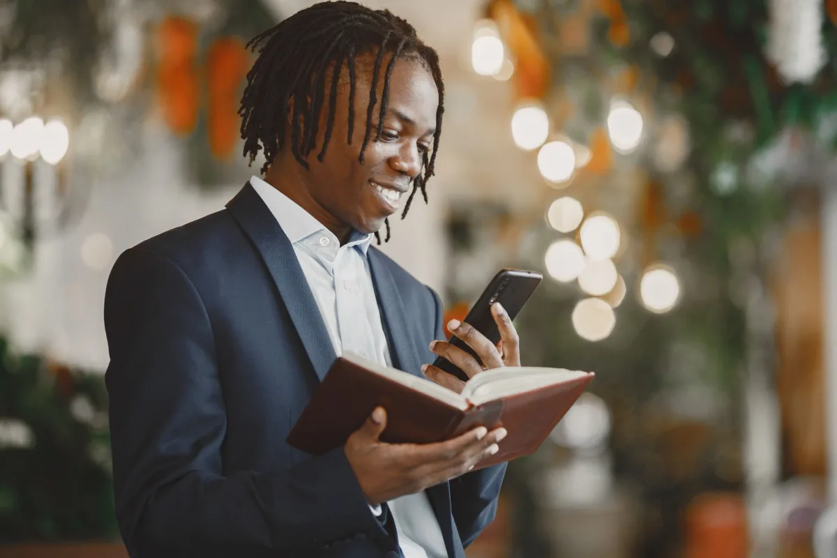Smiling businessman multitasking with book and smartphone in an indoor setting with bokeh lighting. Smiling businessman multitasking with book and smartphone in an indoor setting with bokeh lighting.