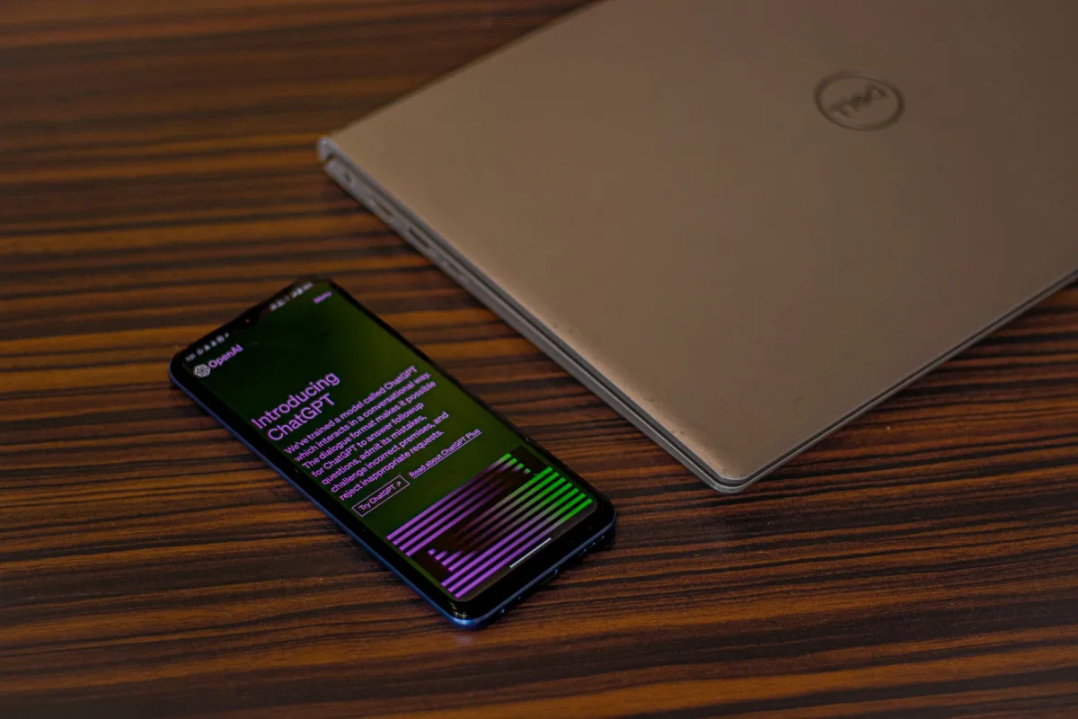 A smartphone displaying ChatGPT app beside a closed laptop on a wooden table. A smartphone displaying ChatGPT app beside a closed laptop on a wooden table.