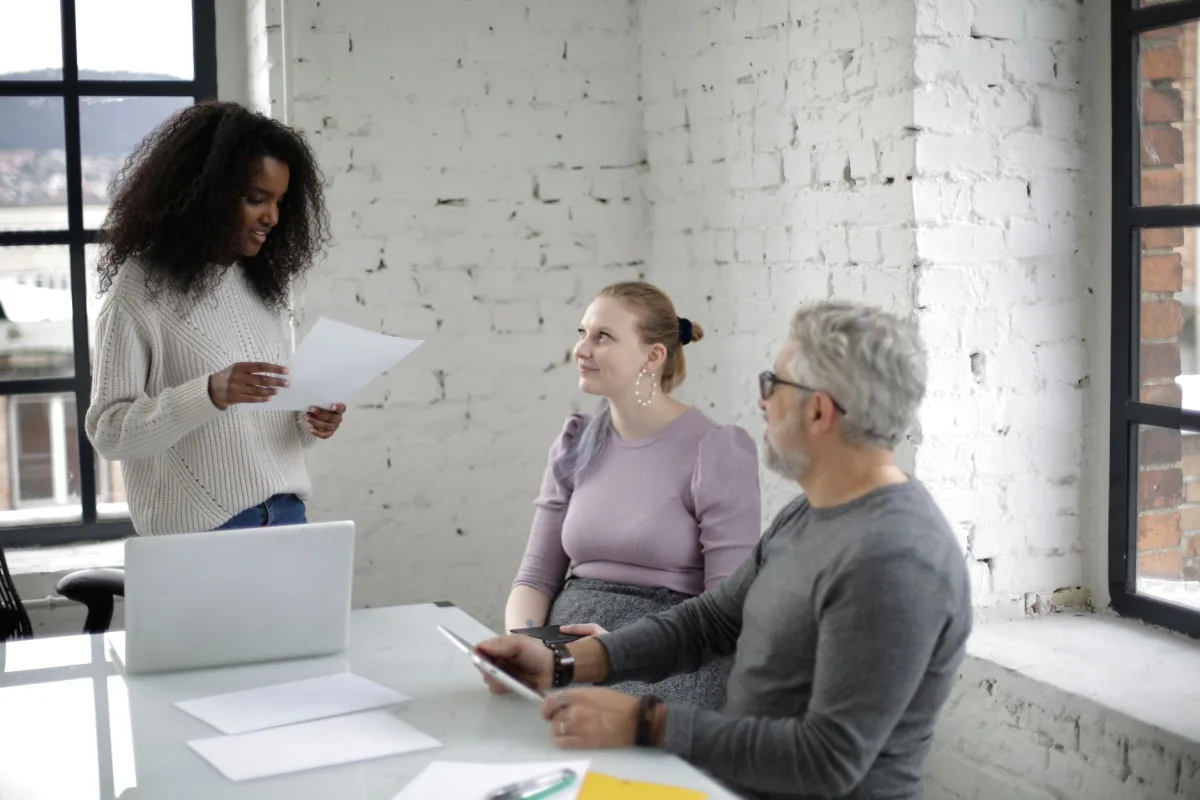 Group of positive multiracial people in casual outfits gathering together and discussing documents while working on tablet and laptop in modern office Group of positive multiracial people in casual outfits gathering together and discussing documents while working on tablet and laptop in modern office