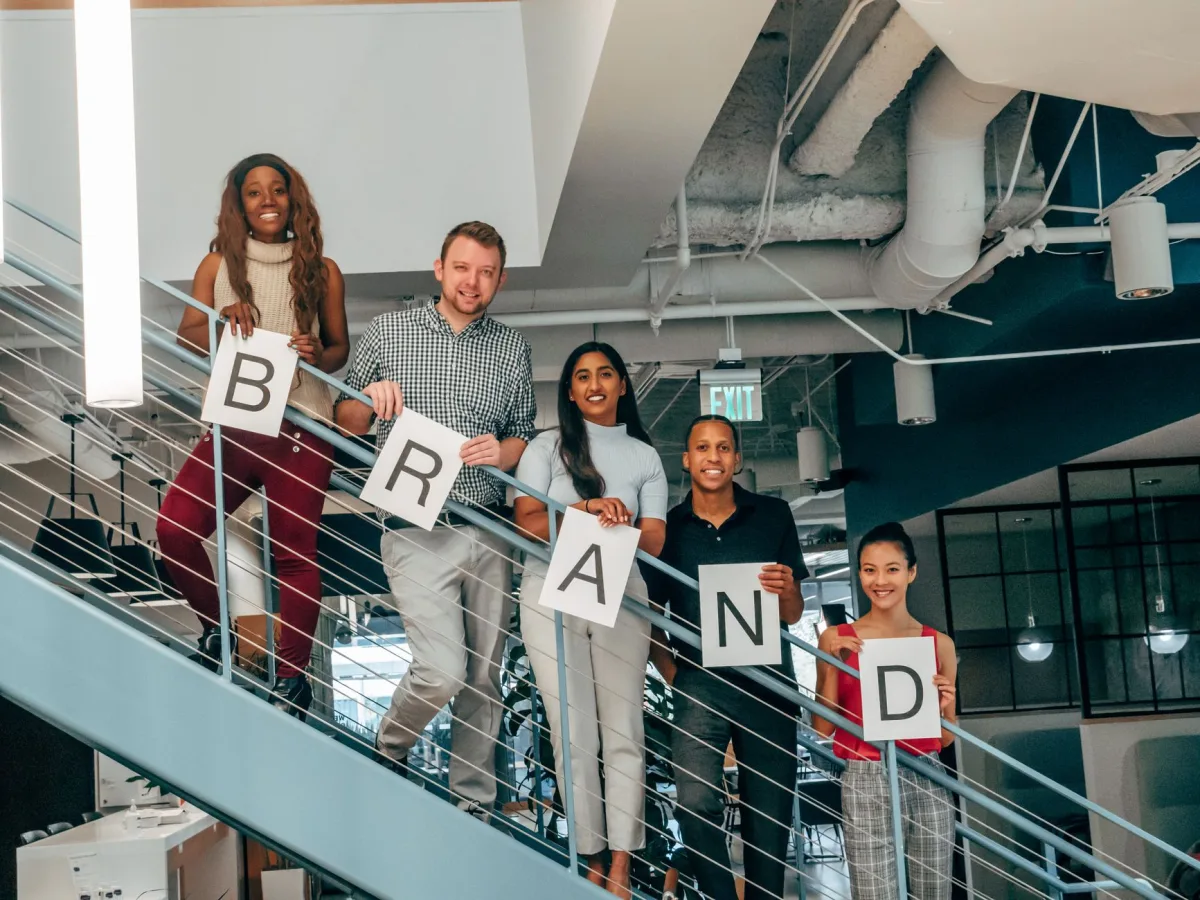 Diverse group of adults smiling and holding 'BRAND' signs on an office staircase. Diverse group of adults smiling and holding 'BRAND' signs on an office staircase.